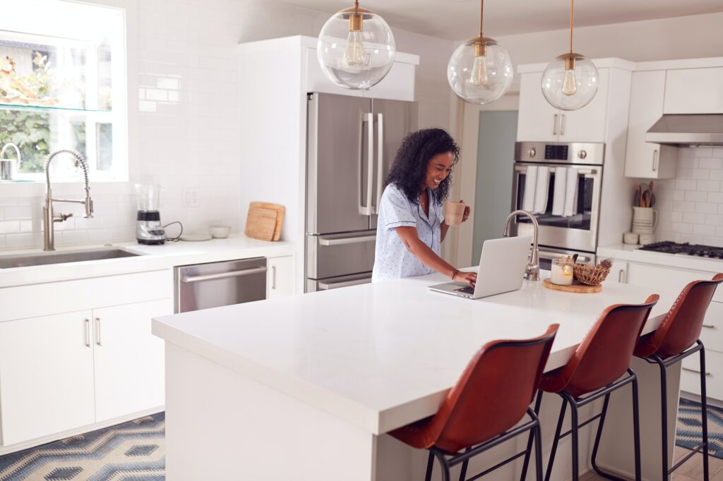 Woman Wearing Pyjamas Standing In Kitchen Working From Home On Laptop